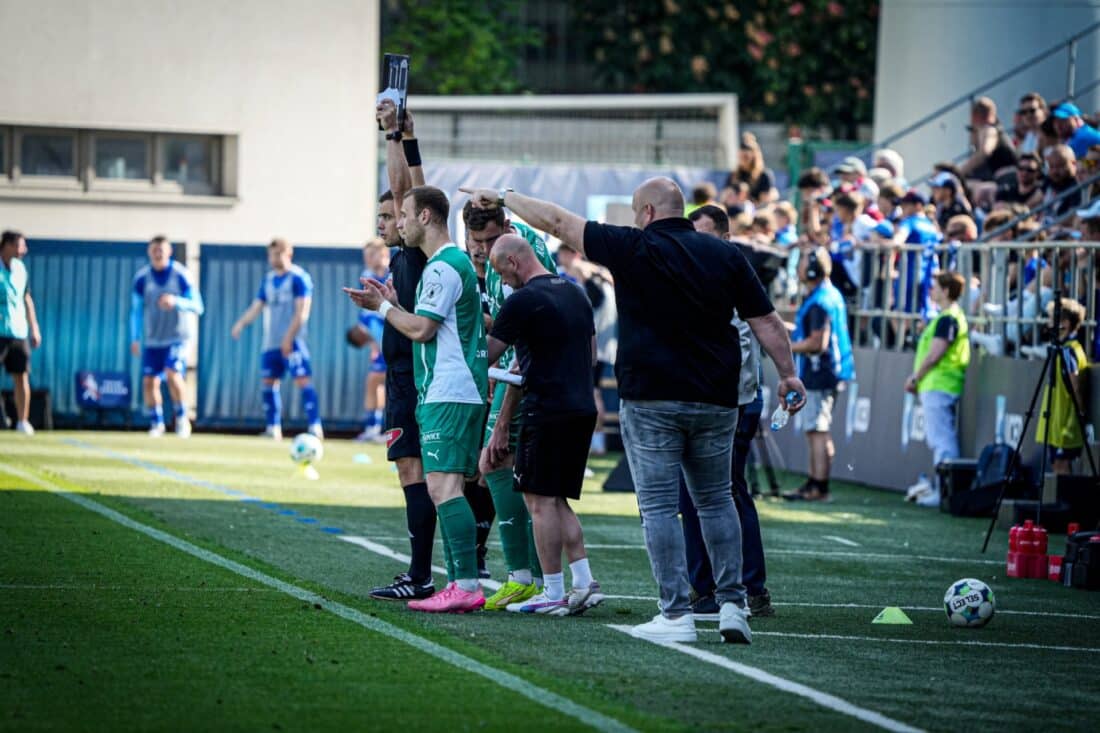 Team members on the sidelines in front of bleachers full of fans at a Bohemians game.