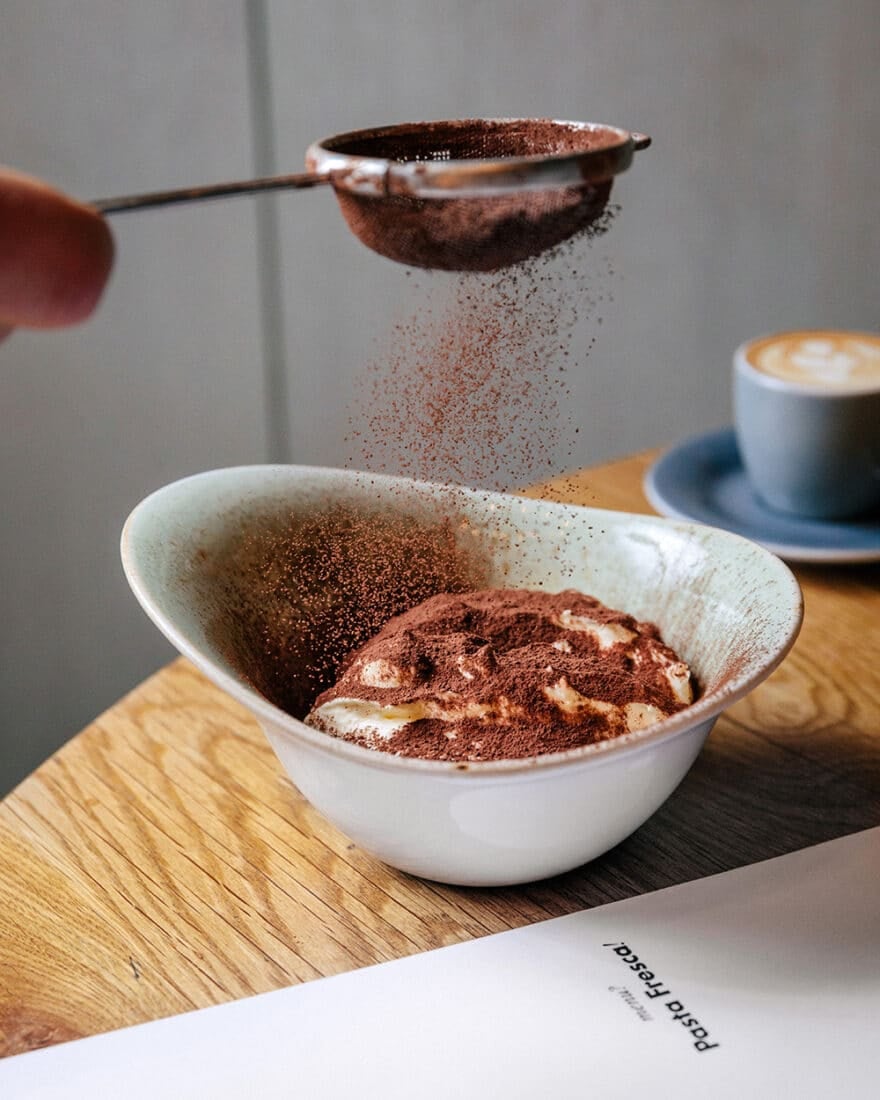 A hand sifting powder over tiramisu at Pasta Fresca, a great Prague alternative to Olive Garden.